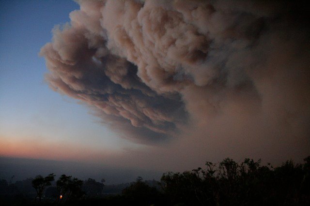 The same smoke column later in the evening, darker underneath, with a faint ember glow at the treeline.