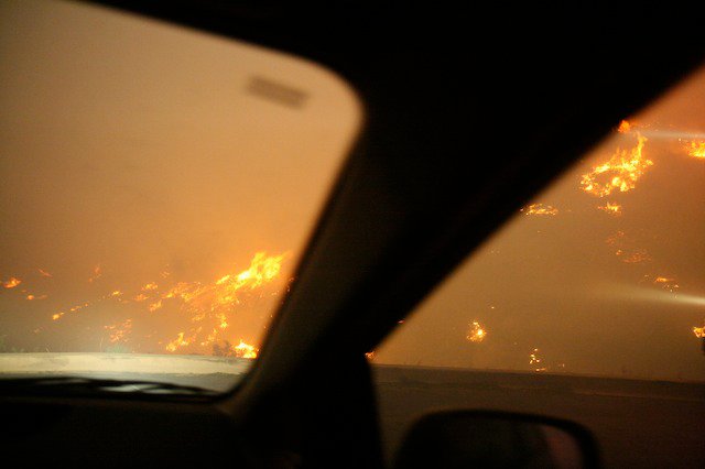 View through a car windshield of an orange flame-field on both sides of the road during evacuation.