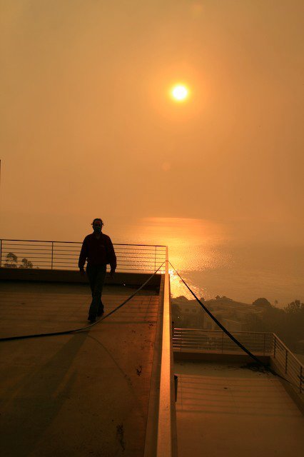 A masked figure on a deck rail as the sun burns dim through wildfire smoke, the Pacific glowing copper below.